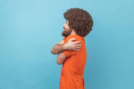 Photo for Side view of selfish narcissistic man with Afro hairstyle embracing himself and smiling with expression of great ego, pleasure and self-esteem. Indoor studio shot isolated on blue background. - Royalty Free Image