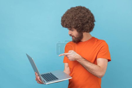 Téléchargez les photos : Portrait d'un homme sérieux avec une coiffure afro portant un T-shirt orange tenant un ordinateur portable et ayant un appel vidéo, pointant du doigt l'affichage du carnet, avertissement. Studio intérieur tourné isolé sur fond bleu. - en image libre de droit