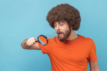 Téléchargez les photos : Portrait d'homme à la coiffure afro en T-shirt orange regardant à travers la loupe, espionnant, découvrant quelque chose, explorant la scène de crime, inspectant. Studio intérieur tourné isolé sur fond bleu - en image libre de droit