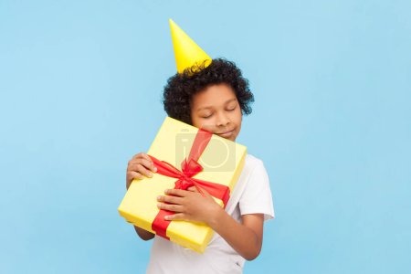 Photo for Portrait of cute little boy with curly hair wearing yellow party cone standing embracing his birthday present enjoying with closed eyes. Indoor studio shot isolated on blue background. - Royalty Free Image