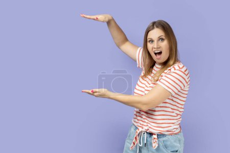Portrait of amazed surprised blond woman wearing striped T-shirt presenting area between hands for advertisement, showing huge size. Indoor studio shot isolated on purple background.