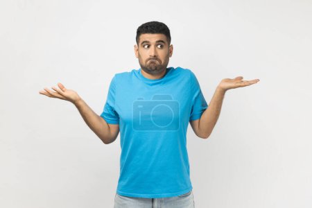 Photo for Portrait of puzzled uncertain unshaven man wearing blue T- shirt standing shrugging shoulders, doesn't know how to make decision. Indoor studio shot isolated on gray background. - Royalty Free Image