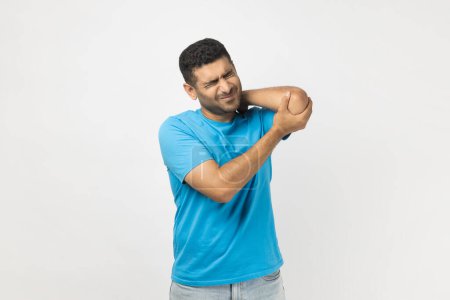 Photo for Portrait of upset unhealthy unshaven man wearing blue T- shirt standing touching his injured elbow, frowning face from terrible pain. Indoor studio shot isolated on gray background. - Royalty Free Image