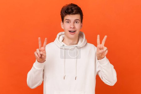 Photo for Teen guy wearing white hoodie shows peace v sign smiles toothily enjoys nice day gestures victory symbol, has good mood, carefree expression. Indoor studio shot isolated on orange background. - Royalty Free Image