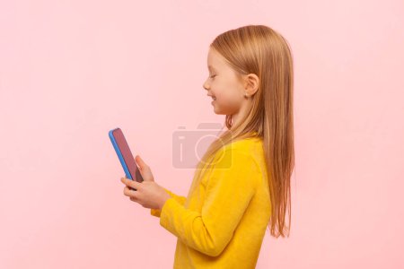 Téléchargez les photos : Vue latérale portrait d'adorable petite fille blonde mignonne utilisant un téléphone portable avec une expression heureuse, jouer à des jeux, profiter, porter un pull jaune. Studio intérieur tourné isolé sur fond rose. - en image libre de droit
