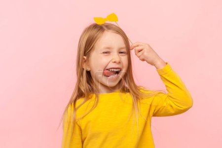 Téléchargez les photos : Portrait of funny positive cheerful blonde little girl holding paper bow above her head, showing tongue out, having fun on party, wearing yellow jumper. Indoor studio shot isolated on pink background. - en image libre de droit