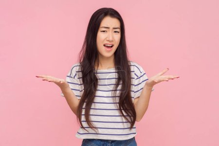 Portrait of shocked unhappy woman with long brunette hair asking what grimacing arguing with somebody, wearing striped T-shirt. Indoor studio shot isolated on pink background.
