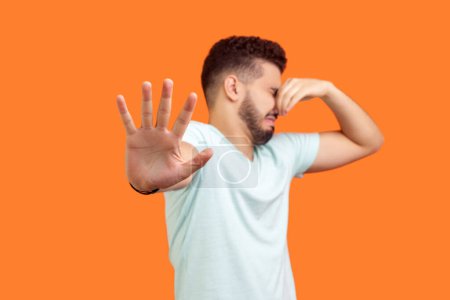 Portrait of displeased young bearded man wearing T-shirt pinching his nose turning away feeling disgust bad smell. Indoor studio shot isolated on orange background.