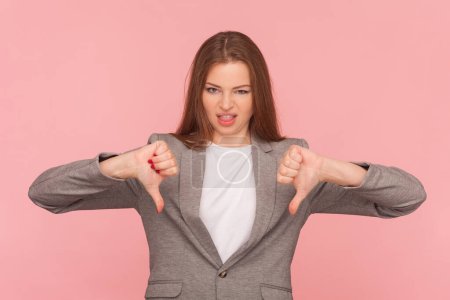 Photo for Portrait of displeased sad woman with brown hair showing dislike gesture, thumbs down,negative feedback, not recommend, wearing business suit. Indoor studio shot isolated on pink background. - Royalty Free Image