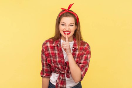 Photo for Portrait of positive cheerful woman keeps finger near red lips keeping secret, smiling with cunning look, wearing checkered shirt and headband. Indoor studio shot isolated on yellow background. - Royalty Free Image