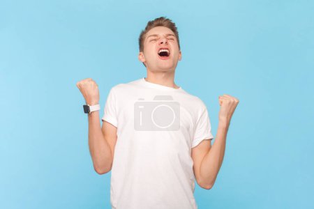 Foto de Retrato de un hombre muy feliz y alegre con una camiseta blanca celebrando sus logros y objetivos, regocijándose por la victoria y el éxito. Estudio interior plano aislado sobre fondo azul - Imagen libre de derechos