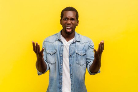 Portrait of angry annoyed man standing with raised arms and frowning face expressing anger and aggression, wearing denim casual shirt. Indoor studio shot isolated on yellow background.