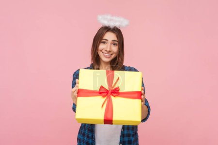 Portrait of cute smiling angelic brown haired woman with nimb over head giving present box, congratulating, offering gift, wearing checkered shirt. Indoor studio shot isolated on pink background.