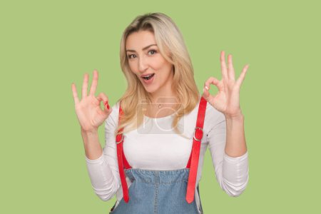 Photo for Portrait of excited positive adult blond woman standing looking at camera, saying everything is ok, showing okay gesture, wearing denim overalls. Indoor studio shot isolated on light green background - Royalty Free Image