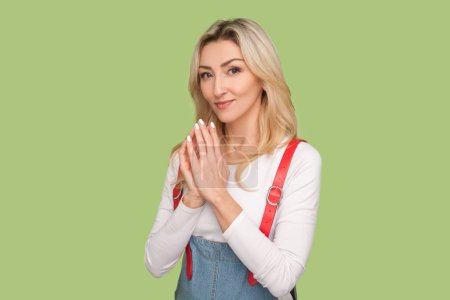 Photo for Portrait of envy cunning beautiful adult blond woman presses fingers together, planning sly prank, looking at camera, wearing denim overalls. Indoor studio shot isolated on light green background - Royalty Free Image