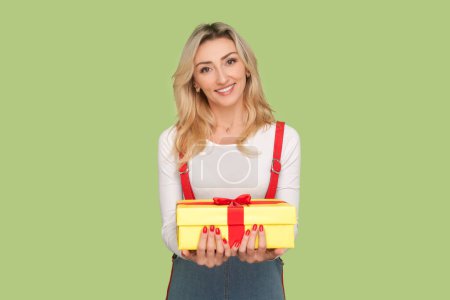Photo for Portrait of satisfied pleased pretty adult blond woman holding present box in hands, congratulating with birthday, wearing denim overalls. Indoor studio shot isolated on light green background - Royalty Free Image