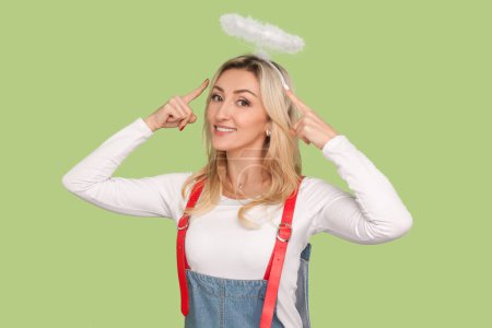 Photo for Portrait of delighted cheerful angelic adult blond woman pointing at her nimb over head, looking smiling at camera, wearing denim overalls. Indoor studio shot isolated on light green background - Royalty Free Image