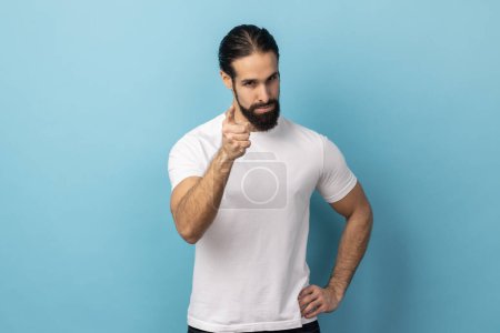 Angry man with beard wearing white T-shirt pointing finger at camera and looking with dissatisfied suspicious expression, warning about troubles. Indoor studio shot isolated on blue background.
