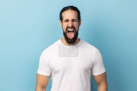 Portrait of playful cheerful man with beard wearing white T-shirt showing tongue, having fun, fooling around, childish manners, keeps eyes closed. Indoor studio shot isolated on blue background.
