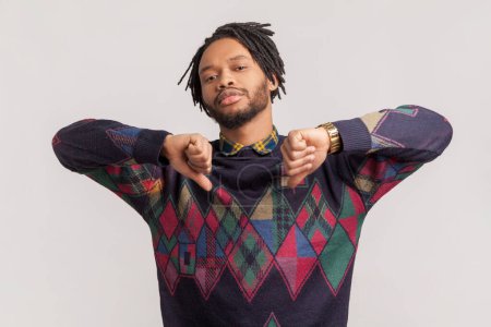 Photo for Portrait of sad disappointed attractive african-american man with dreadlocks and beard showing dislike gesture with both hands, thumbs down. Indoor studio shot isolated on gray background. - Royalty Free Image