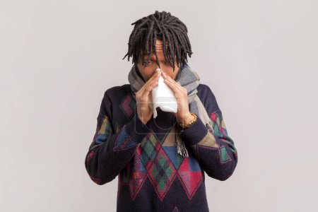 Portrait of sick unhealthy african-american man with dreadlocks and beard standing wrapped in scarf, suffering runny nose, flu symptoms. Indoor studio shot isolated on gray background.
