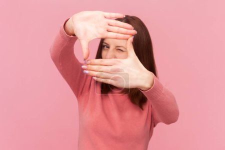 Photo for Focused attractive woman photographer with brown hair standing looking through frame made with her fingers, wearing rose turtleneck. Indoor studio shot isolated on pink background - Royalty Free Image