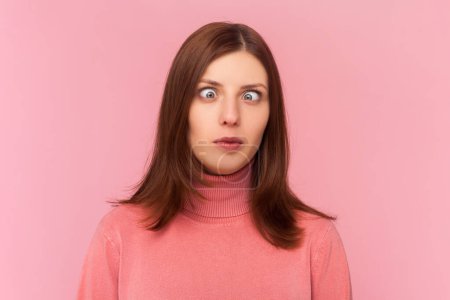 Attractive funny silly woman with brown hair with cross eyed, has stupid dumb face, awkward confused comical expression, wearing rose turtleneck. Indoor studio shot isolated on pink background