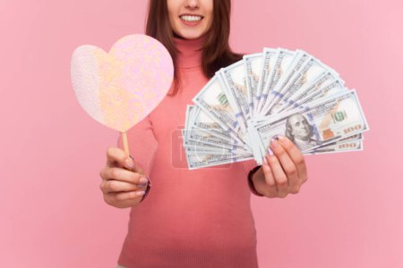 Téléchargez les photos : Unknown smiling woman with brown hair holding little pink heart on stick and fan of dollar banknotes, love to money, wearing rose turtleneck. Indoor studio shot isolated on pink background - en image libre de droit