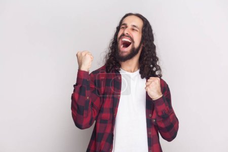 Emotional bearded man raises clenched fists, exclaims with excitement, rejoices sweet success, feels taste of victory, shouts for favorite team. Indoor studio shot isolated on gray background.