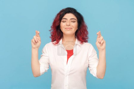 Photo for Portrait of hopeful smiling adult woman with fancy red hair crossing fingers, making wish, hopes for good luck, wearing white shirt. Indoor studio shot isolated on blue background. - Royalty Free Image