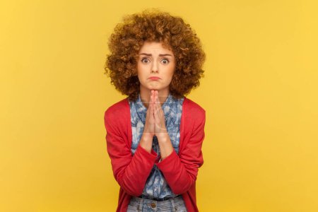 Porträt einer flehenden Frau mit Afro-Frisur, die in Gebetsgeste gefaltete Hände hält, um Hilfe bittet und die Handflächen zusammenpresst. Indoor Studio aufgenommen isoliert auf gelbem Hintergrund.