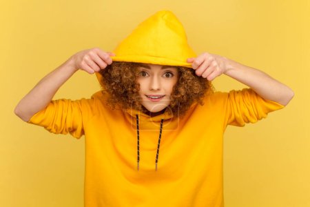 Photo for Portrait of positive woman with Afro hairstyle putting on her hood, looking at camera with funny face, wearing casual style hoodie. Indoor studio shot isolated on yellow background. - Royalty Free Image