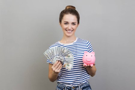 Photo for Portrait of delighted satisfied woman wearing striped T-shirt holding fan of dollar bills and piggy bank, savings, growth of deposit. Indoor studio shot isolated on gray background. - Royalty Free Image
