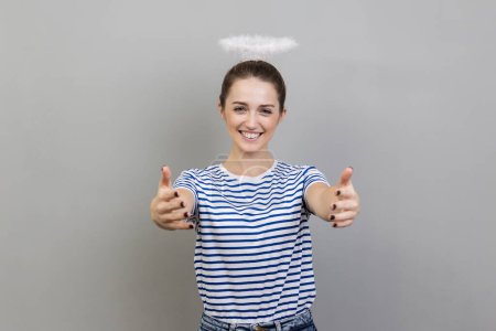 Portrait of woman wearing striped T-shirt and with nimbus over her head stretching arms to camera with kind friendly smile, going to embrace, share love. Indoor studio shot isolated on gray background