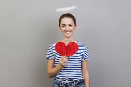 Portrait of romantic smiling woman wearing striped T-shirt and with nimbus over her head holding red heart, looking at camera with toothy smile. Indoor studio shot isolated on gray background.