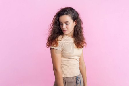 Photo for Portrait of cute pretty teenage girl with wavy hair in beige T- shirt standing looking away with positive facial expression. Indoor studio shot isolated on pink background. - Royalty Free Image