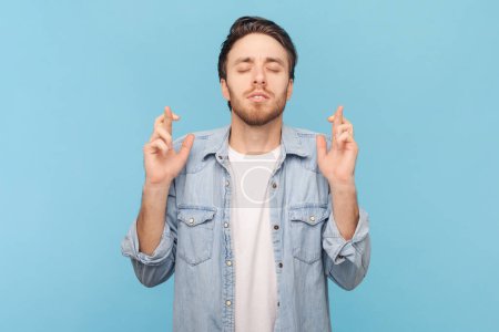 Portrait of man with bristle standing with crossed fingers for good luck, heartily making wish, praying and hoping for miracle, wearing denim shirt. Indoor shot isolated on blue background.