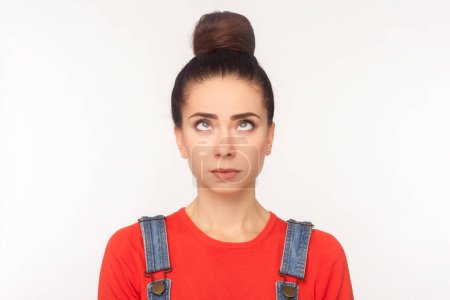 Portrait of funny childish playful woman with hair bun looking with silly comedian face having fun alone, wearing denim overalls. Indoor studio shot isolated on white background