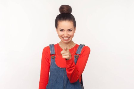 Portrait of joyous pretty attractive woman with hair bun pointing to camera, expresses choice, smiles broadly, wearing denim overalls. Indoor studio shot isolated on white background