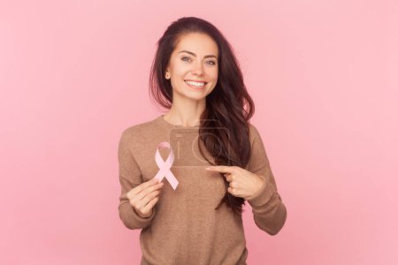 Photo for Portrait of adorable smiling woman with wavy hair, holding pointing at pink ribbon, symbol female health, wearing wearing brown pullover. Indoor studio shot isolated on pink background - Royalty Free Image