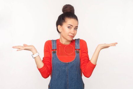 Photo for Portrait of uncertain puzzled woman with hair bun shrugging shoulders, dosen't know answer the question, wearing denim overalls. Indoor studio shot isolated on white background - Royalty Free Image