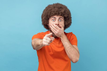 Porträt eines schockierten Mannes mit Afro-Frisur, der ein orangefarbenes T-Shirt trägt und mit großen Augen und offenem Mund aussieht, mit dem Finger in die Kamera zeigt und einen verdächtigt. Indoor Studio aufgenommen isoliert auf blauem Hintergrund.