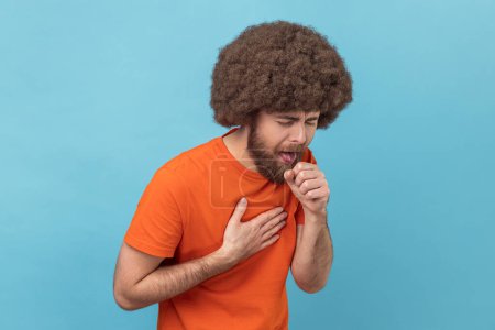 Portrait of sick man with Afro hairstyle wearing orange T-shirt coughing, catches cold, having high temperature, having influenza symptom. Indoor studio shot isolated on blue background.