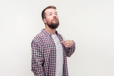 Portrait of confident egoistic bearded man standing pointing at himself looks selfish having proud expression, wearing casual checkered shirt. Indoor studio shot isolated on gray background.