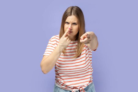 Photo for Suspicious blond woman in T-shirt pointing to camera, touching nose, doing liar gesture, body language symbol of cheats, falsehood and deception. Indoor studio shot isolated on purple background. - Royalty Free Image