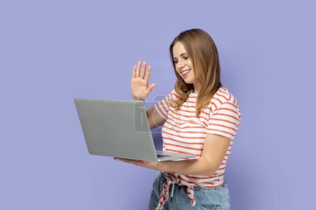 Photo for Portrait of friendly woman wearing striped T-shirt working on laptop, looking at screen and waving hand, saying hello or bye, broadcasting livestream. Indoor studio shot isolated on purple background. - Royalty Free Image