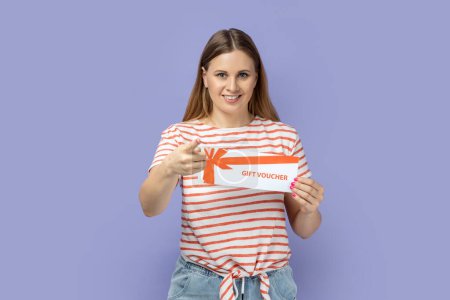 Photo for Portrait of positive smiling blond woman wearing striped T-shirt holding gift voucher and pointing finger to camera, choosing you as a winner. Indoor studio shot isolated on purple background. - Royalty Free Image