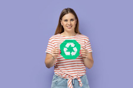 Photo for Portrait of cheerful charming cute blond woman wearing striped T-shirt holding green recycling sign, call on to take care about environment. Indoor studio shot isolated on purple background. - Royalty Free Image
