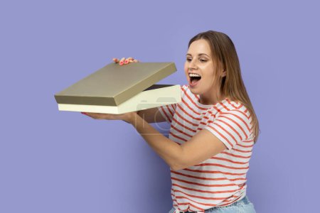 Photo for Portrait of excited amazed blond woman wearing striped T-shirt standing holding present box, looking inside gift with excitement. Indoor studio shot isolated on purple background. - Royalty Free Image