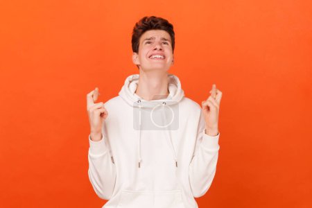 Portrait of hopeful young man wearing white hoodie crossed finger with hope, hopes dreams come true, believes in something important. Indoor studio shot isolated on orange background.